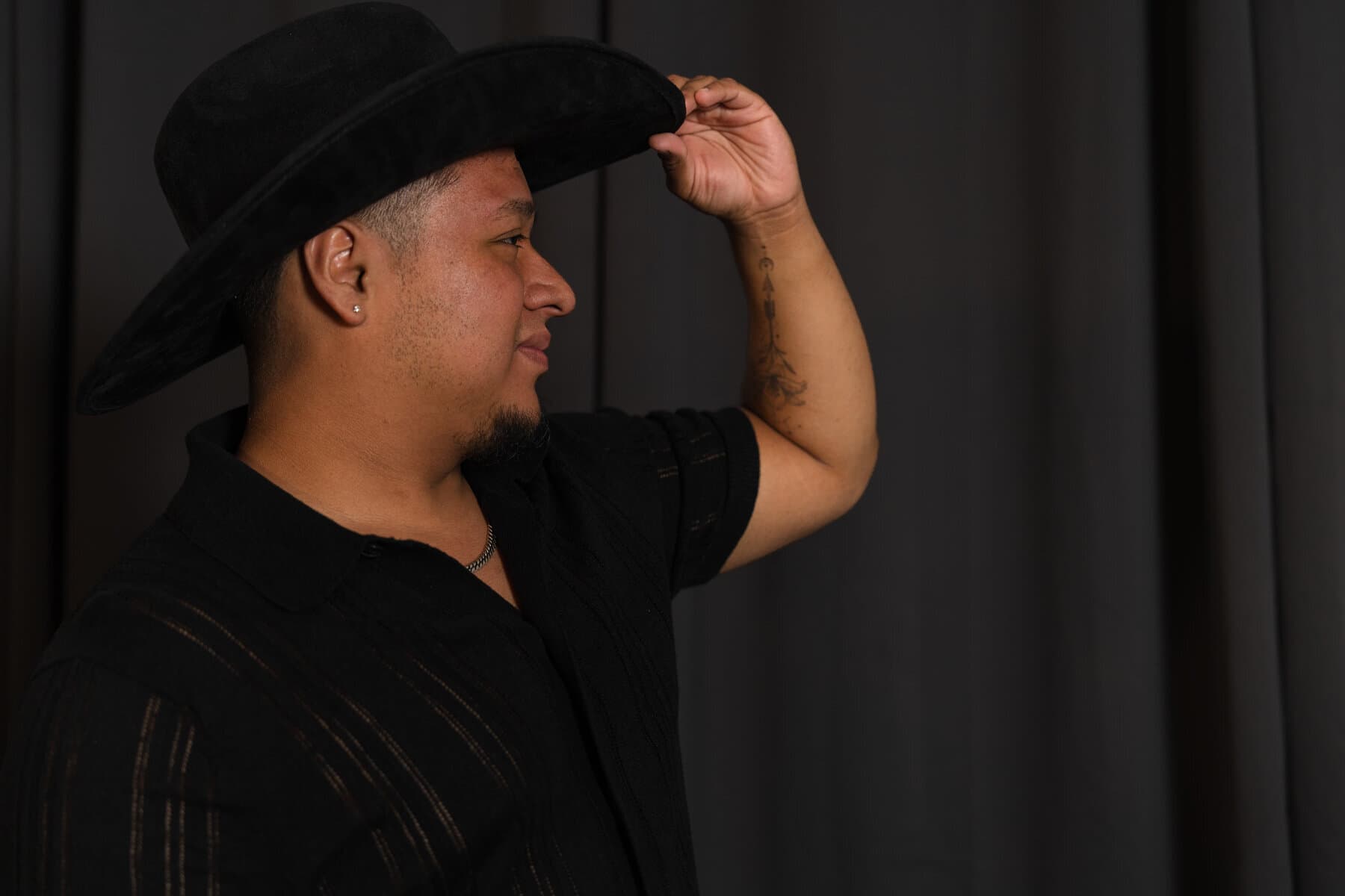 Portrait of a man tipping his cowboy hat, arm tattoo visible, studio backdrop